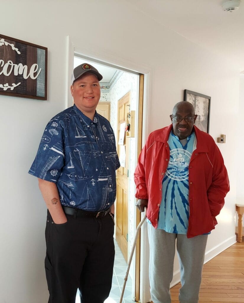 To men standing together in a hallway. A welcome sign hangs on the wall behind them.