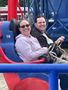 two people are smiling sitting in a red and blue seat holding onto a safety bar in front of them. they waiting for their ride at busch gardens to start.