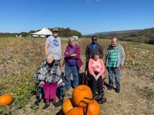 An inclusive group of people are pictured in the field of a local pumpkin patch with pumpkins they are purchasing taking home to decorate.
