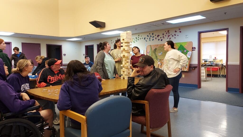 Volunteers playing games at Pleasant View. They have built the Jenga Tower very high. A man in the foreground looks back over his shoulder as if he is nervous it will fall.