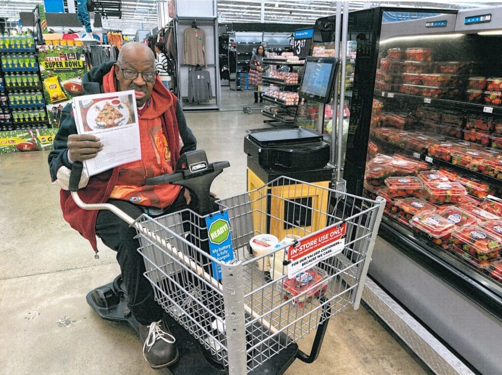 Dennis is sitting in a motorized shopping cart shopping holding up a recipe he is shopping for.